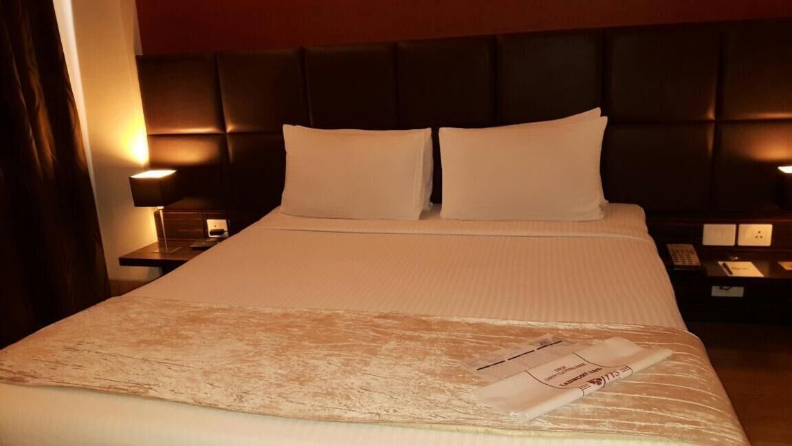 A hotel room with a large bed, two pillows, a headboard, bedside tables with lamps, and a nightstand with a phone and a remote control.