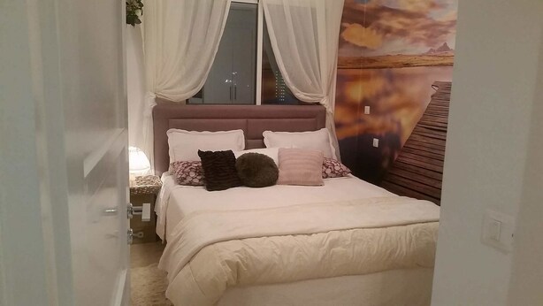 A bedroom with a bed, white curtains, a nightstand, a lamp, and a wall with a landscape painting. - Appartement Haut Standing Lac 2 (Tunis)