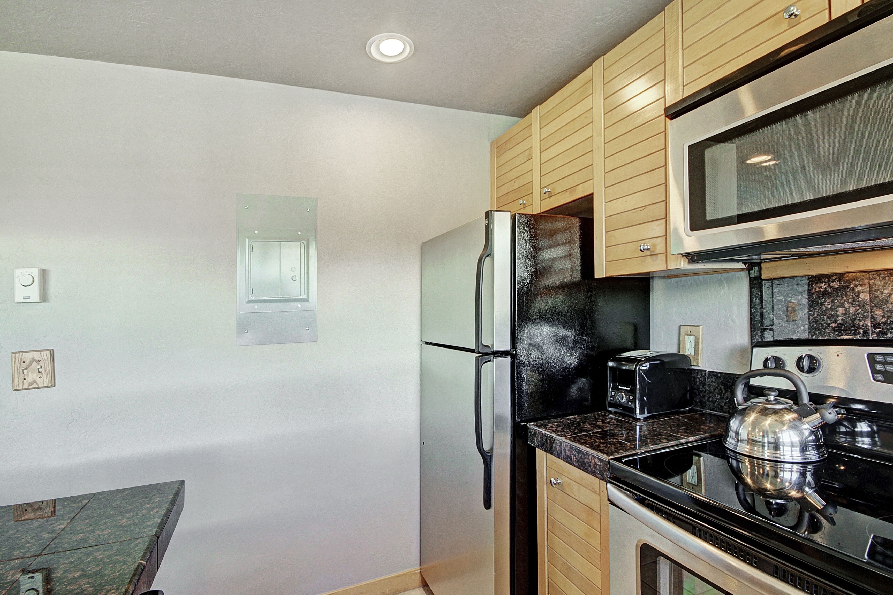 A kitchen with wooden cabinets, a stainless steel refrigerator, a microwave, a stove, and a kettle.
