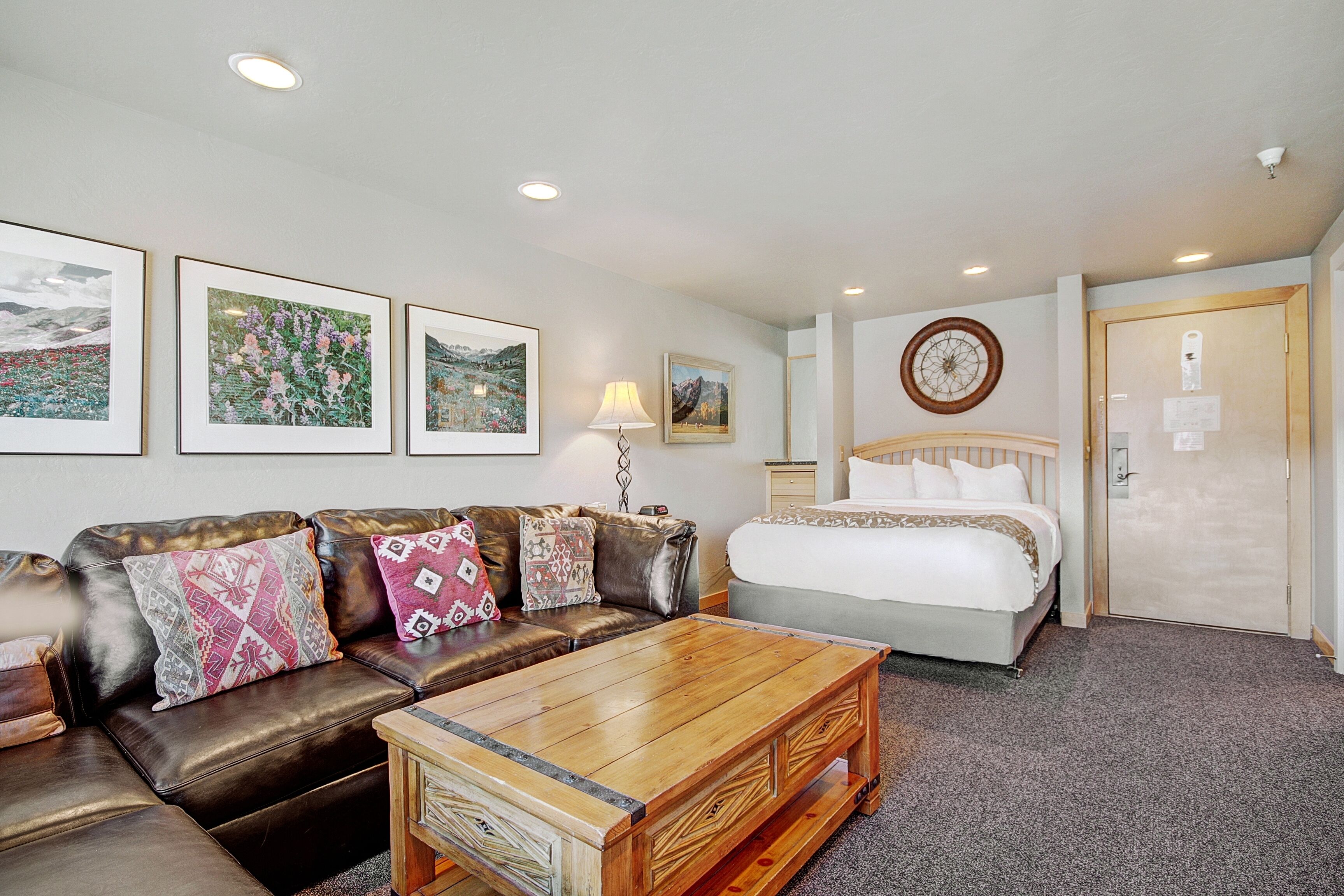 A hotel room with a bed, a leather sofa with patterned cushions, a wooden coffee table, and framed pictures on the wall.