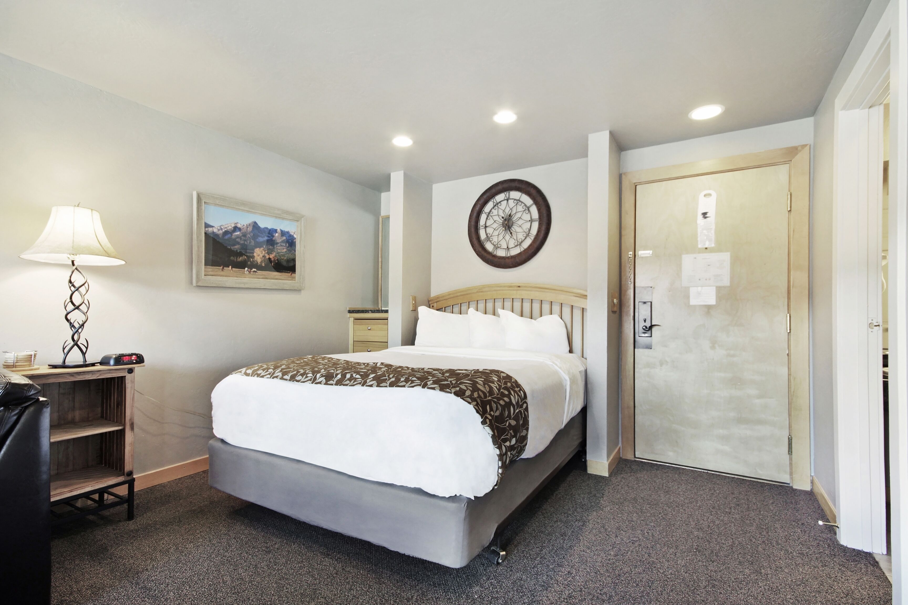 A hotel room with a bed, a desk with a lamp, a clock on the wall, and a door.