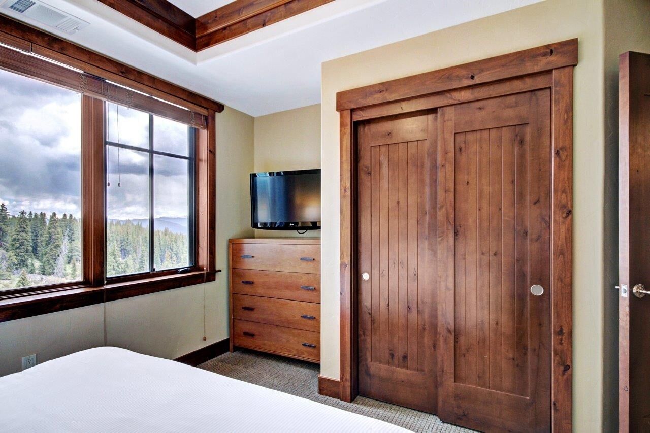 Une chambre avec une grande fenêtre, une armoire en bois, un téléviseur à écran plat et une porte en bois.