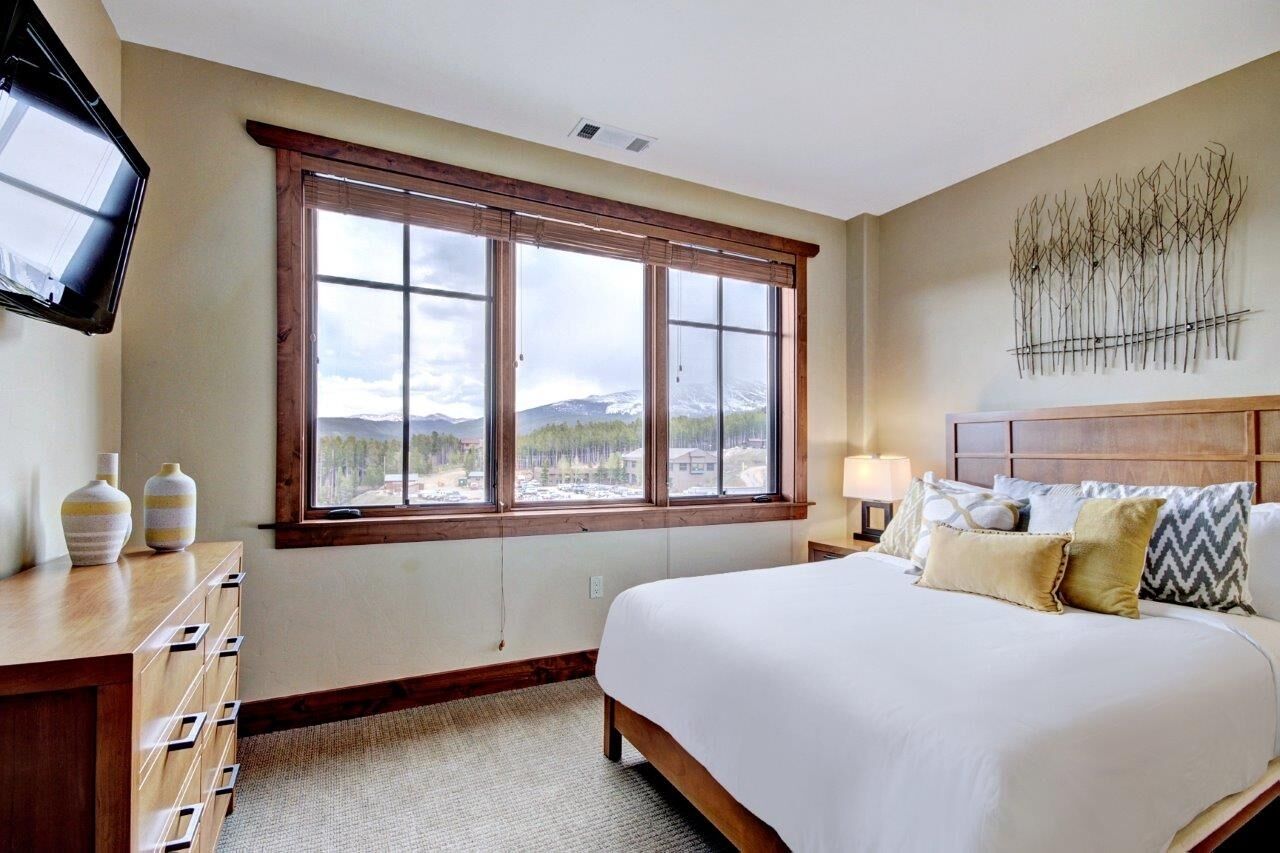 A bedroom with a large window, a bed, a wooden dresser, and a wall-mounted TV.