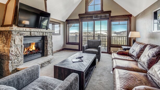 A cozy living room with a stone fireplace, comfortable seating, and large windows offering a view of the mountains. - Premier 2br  At The Base Of Peak 7 2 Bedroom Condo by RedAwning (Breckenridge)