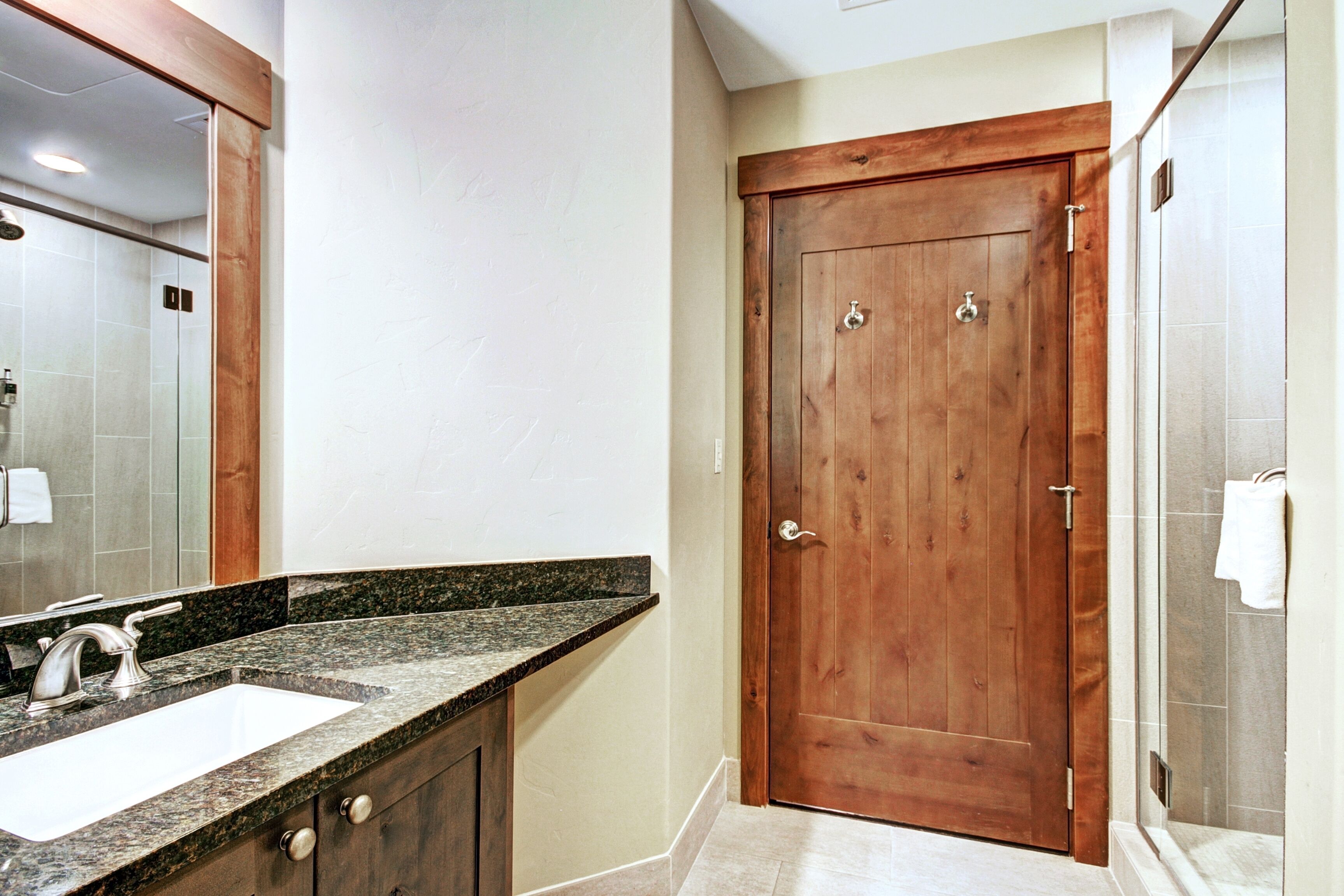 A bathroom with a wooden door, a sink with a granite countertop, and a shower area.