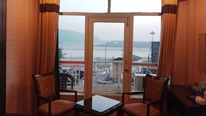 A room with two wooden chairs and a small table, a large window with curtains, and a view of a river and distant buildings. - Paradise Ganga - A River Side Hotel (Rishikesh)
