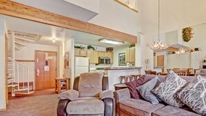A spacious living area with a high ceiling, wooden beams, a kitchen with wooden cabinets, a dining area with a chandelier, and a staircase leading to an upper level.