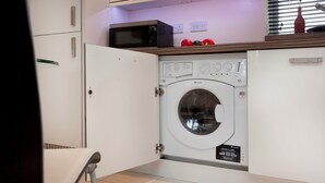 A compact laundry room with a front-loading washing machine, a microwave, and a small countertop with decorative items. - The Coach House (Derby)