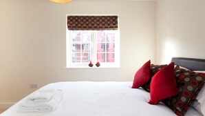 A neatly made bed with white linens and red and brown patterned pillows, a window with a polka dot valance, and a hanging light fixture. - The Coach House (Derby)