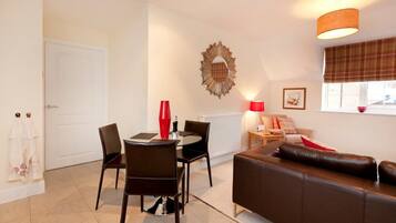 A modern living room with a brown leather sofa, a dining table with chairs, a red vase, and a wall-mounted mirror.