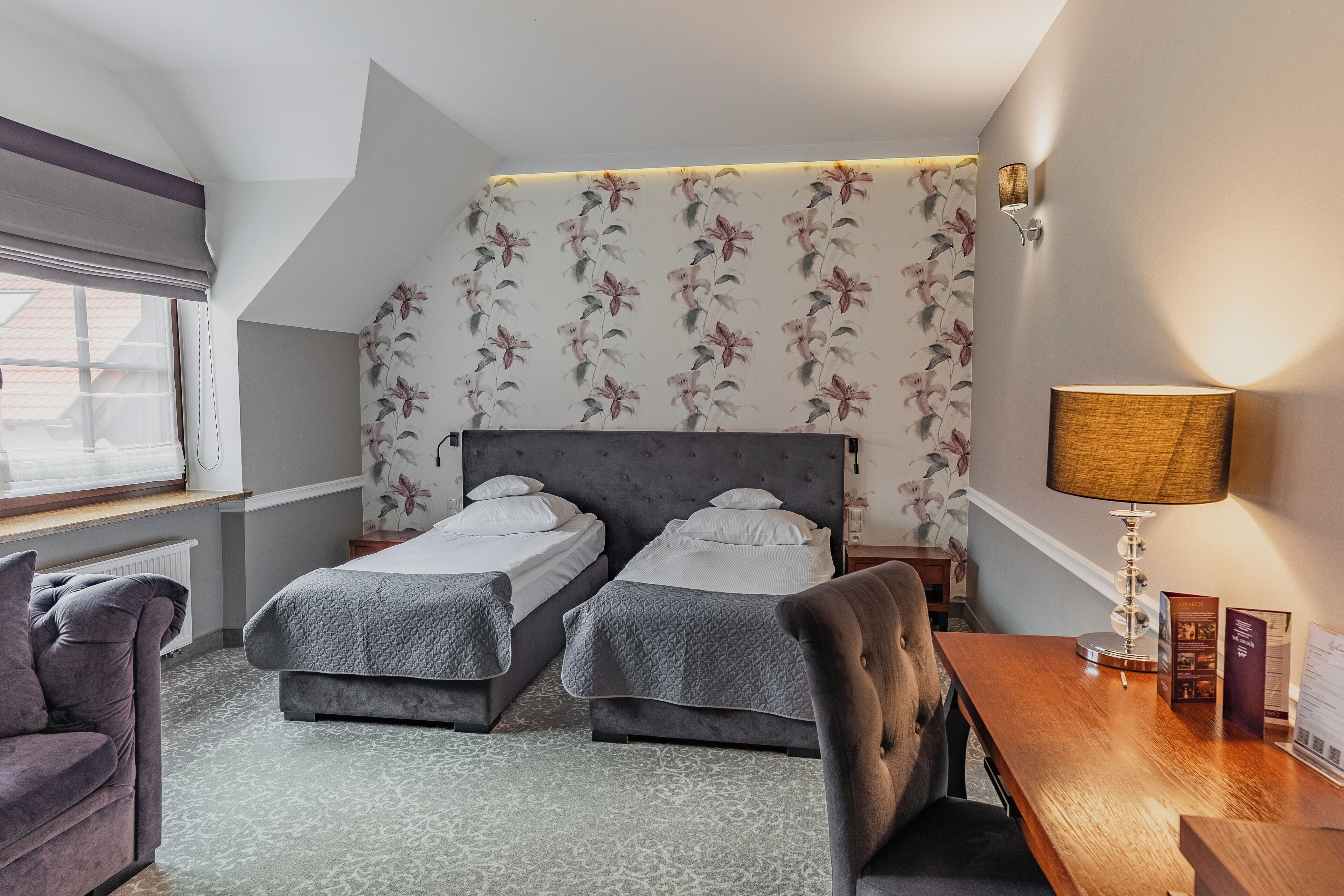 A hotel room with two beds, a desk, a chair, a lamp, and a window with a view.