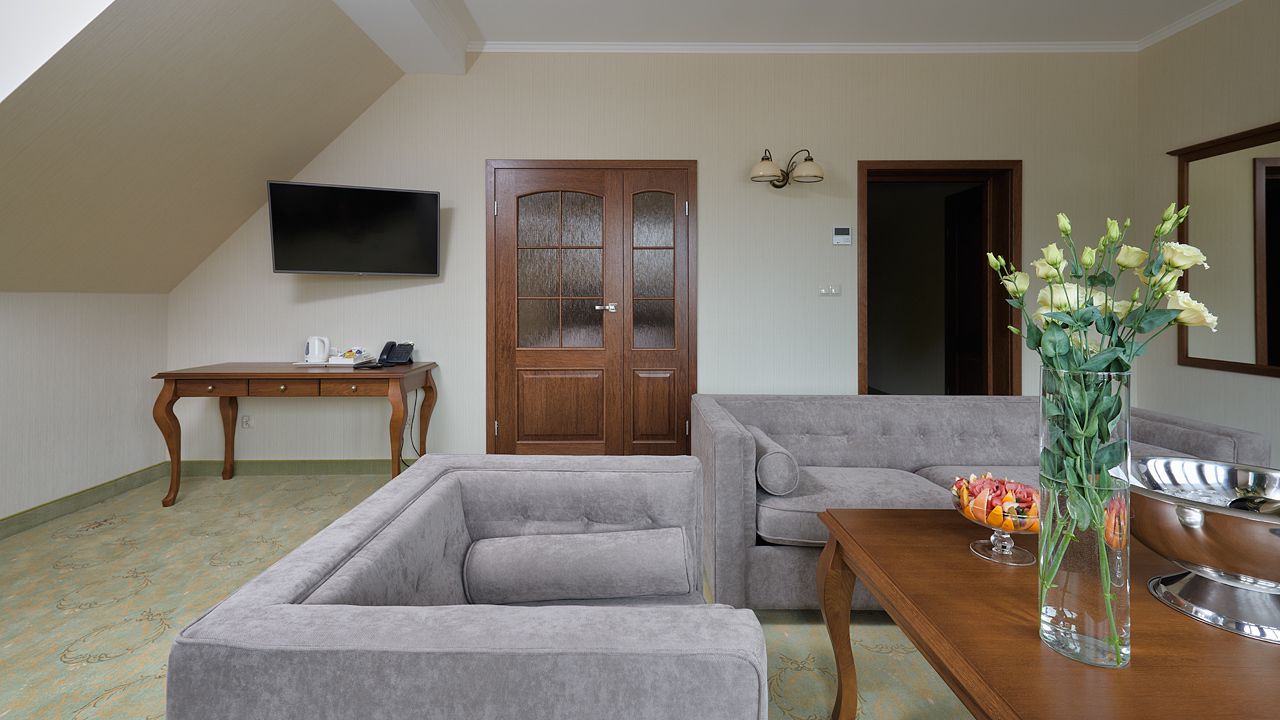 Un salón con un sofá gris, una mesa de centro de madera, un televisor de pantalla plana montado en la pared, una puerta de madera y un jarrón con flores sobre una mesita auxiliar.