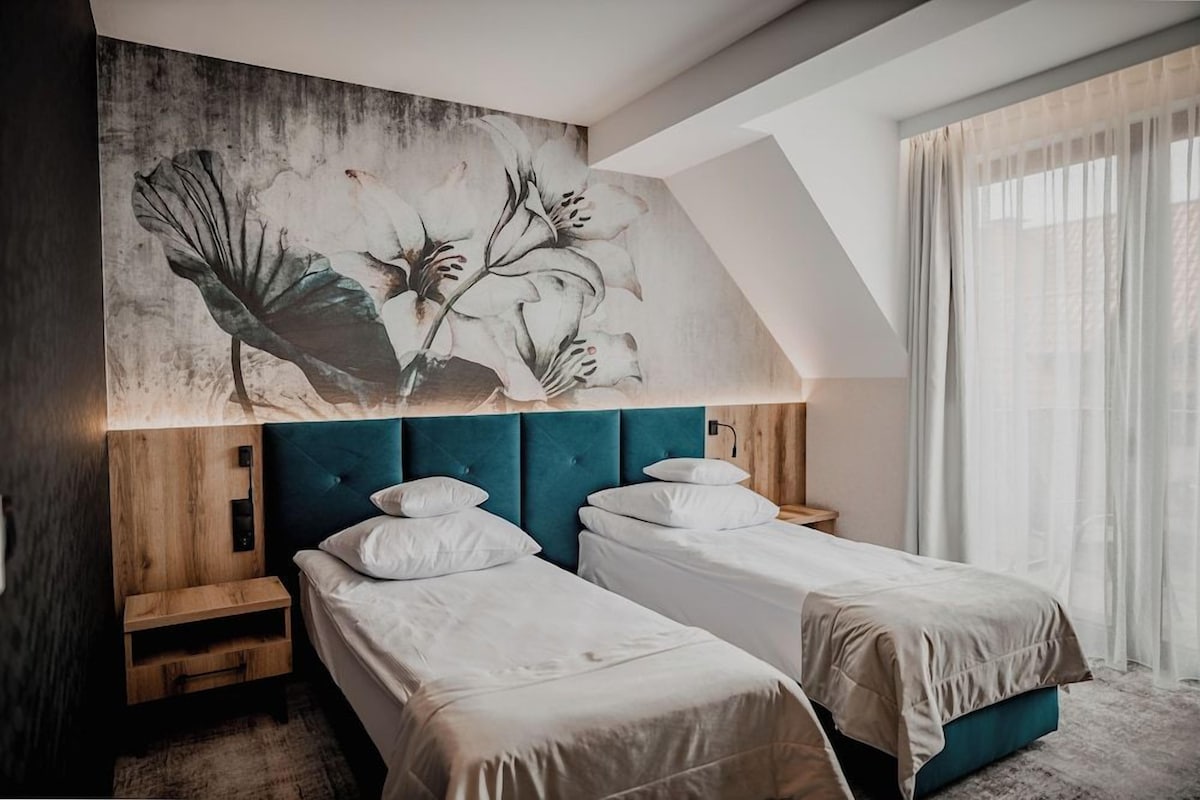 A bedroom with two beds, a wooden headboard, a large floral artwork, and a window with sheer curtains.