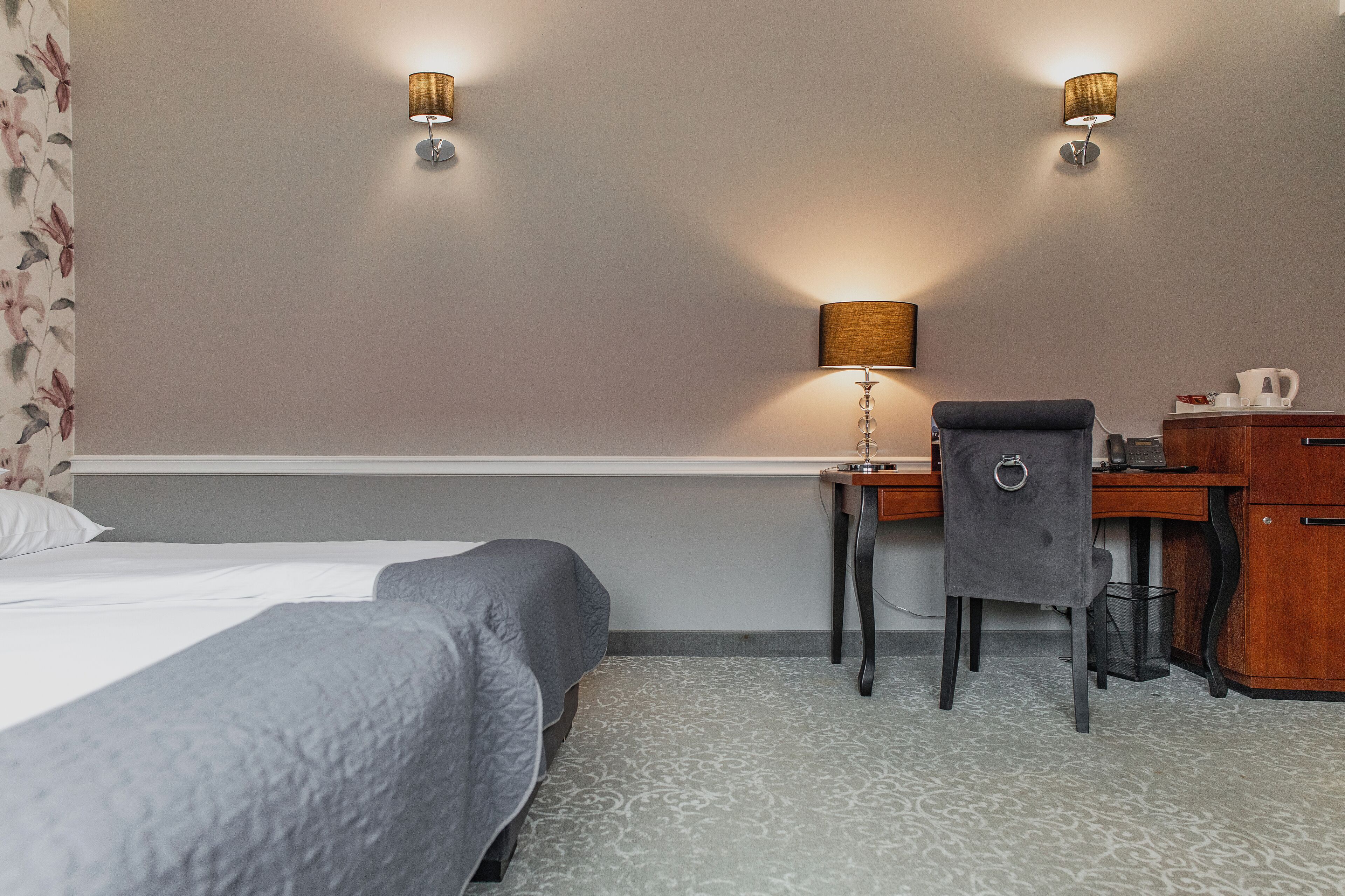 A hotel room with two beds, a desk with a chair, a lamp, and a small table with a coffee maker.