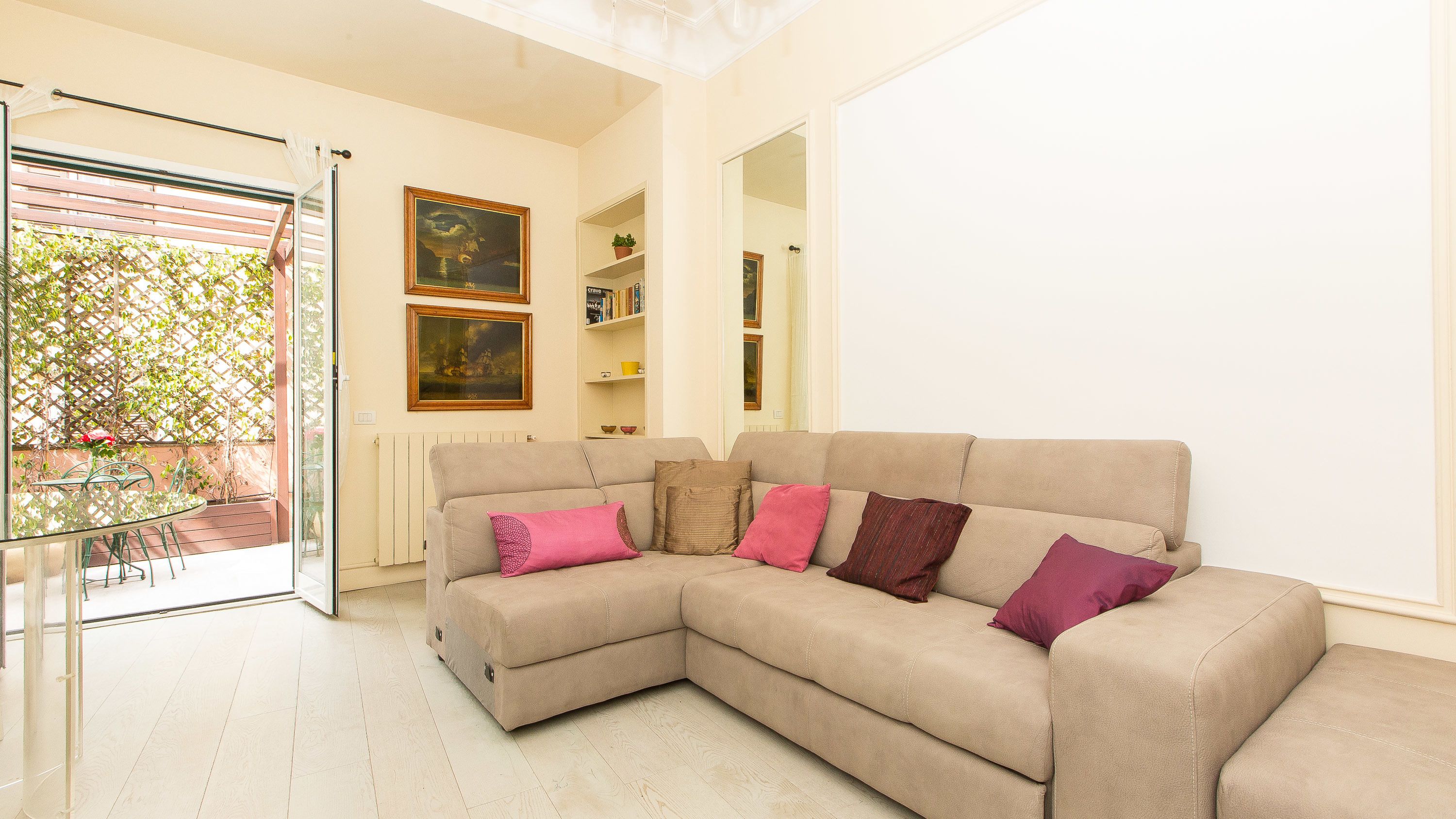 A living room with a beige sectional sofa, a glass table, and chairs, a radiator, and a door leading to a balcony with a lattice and potted plants.