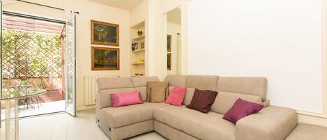 A living room with a beige sectional sofa, a glass table, and chairs, a radiator, and a door leading to a balcony with a lattice and potted plants.