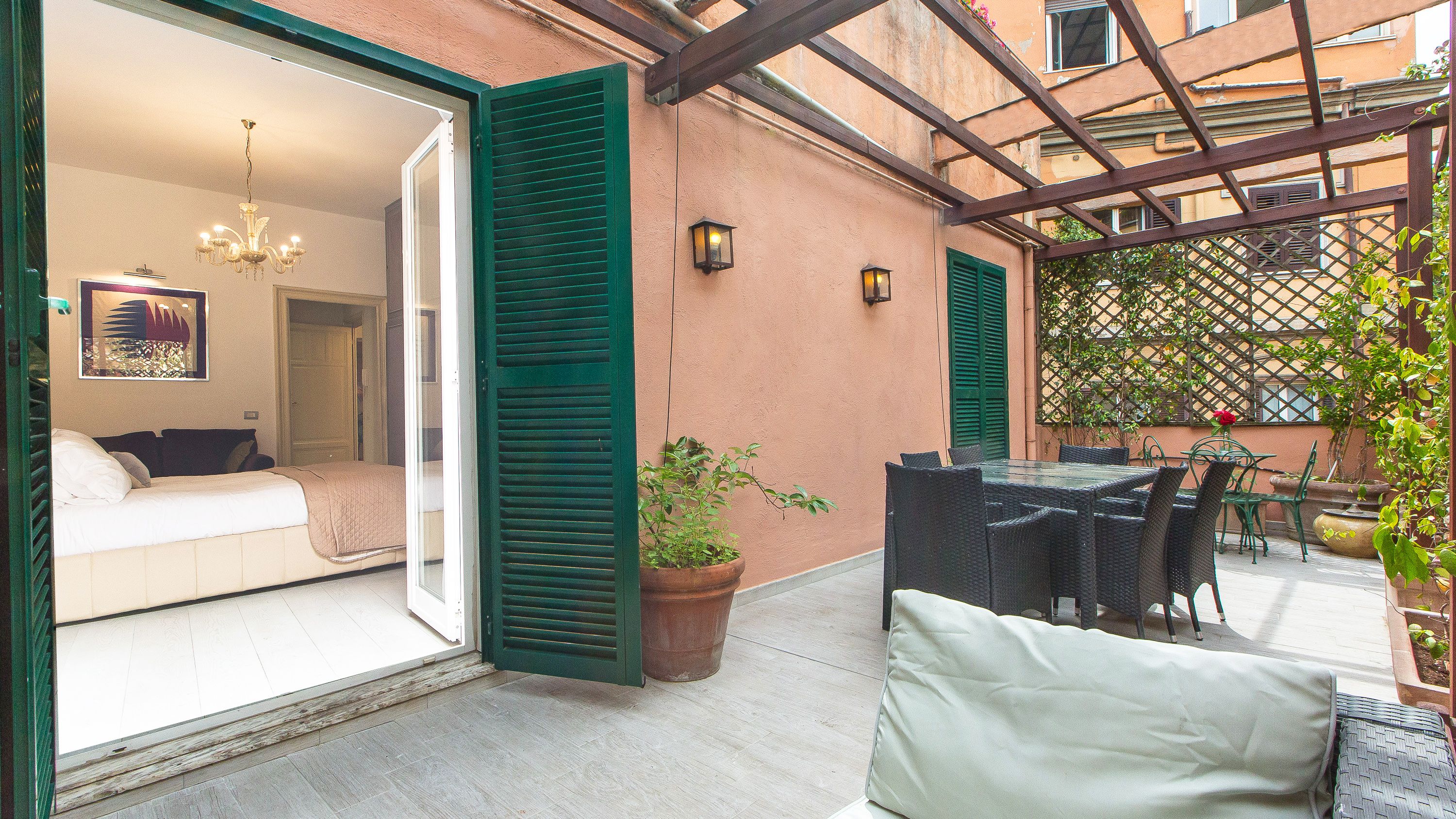 A cozy outdoor patio with a glass table and chairs, a potted plant, and a bed with green shutters. - Rental in Rome Augustus Terrace Deluxe (Rome)