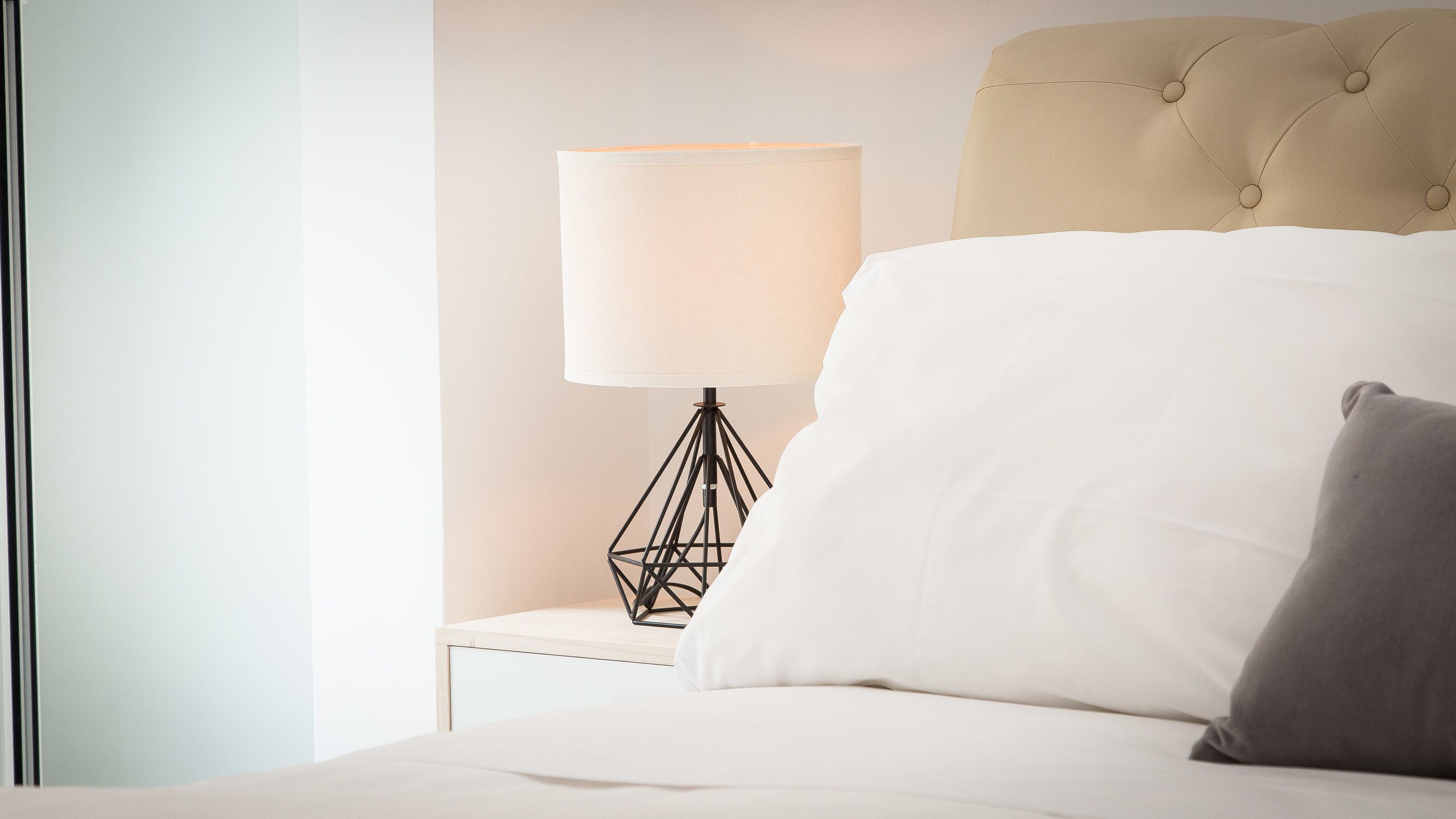 A neatly made bed with white and gray pillows, a beige headboard, and a modern floor lamp with a geometric base.