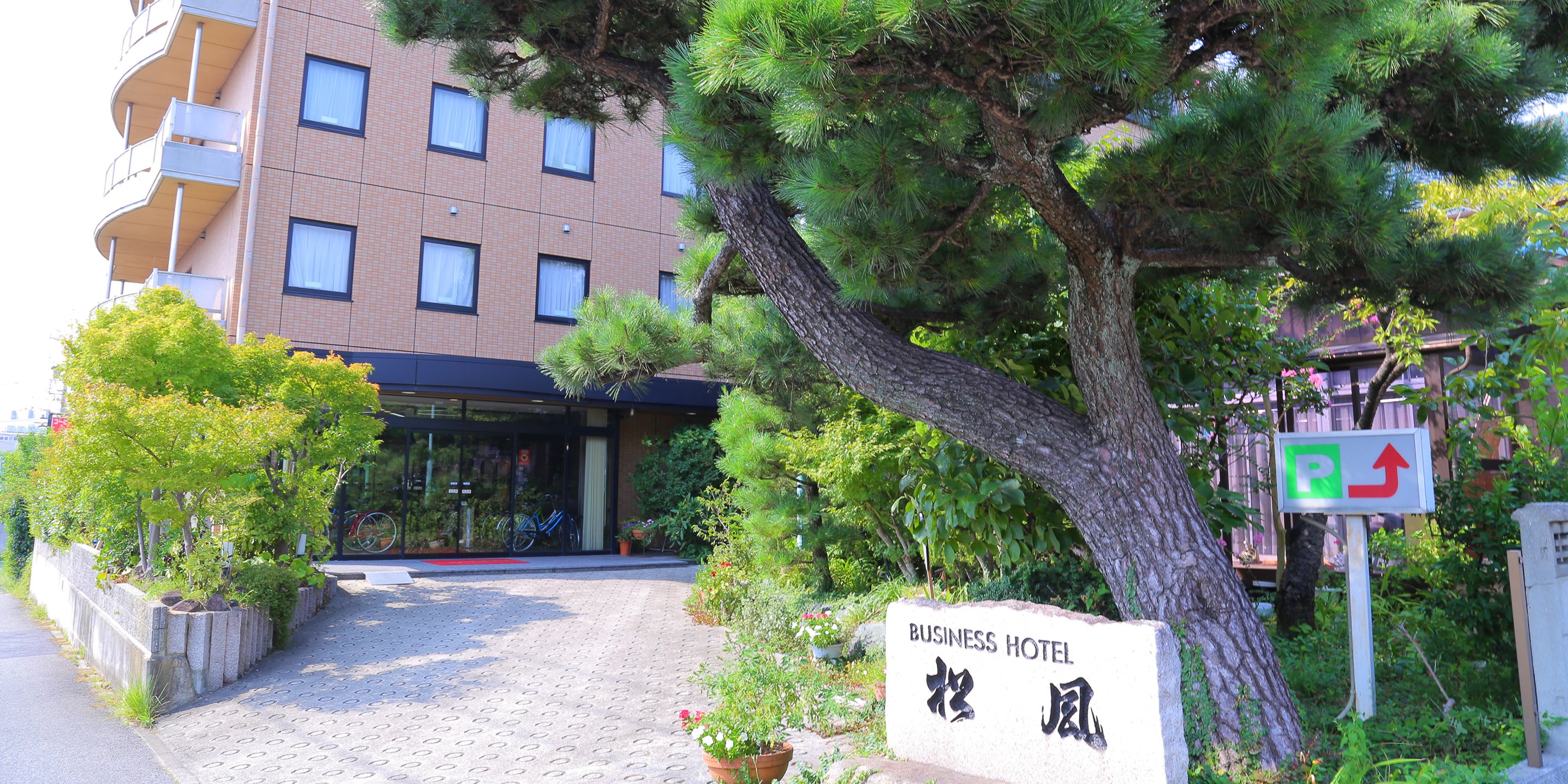 A business hotel entrance with a paved pathway leading to the building, surrounded by greenery and a sign indicating a parking area.