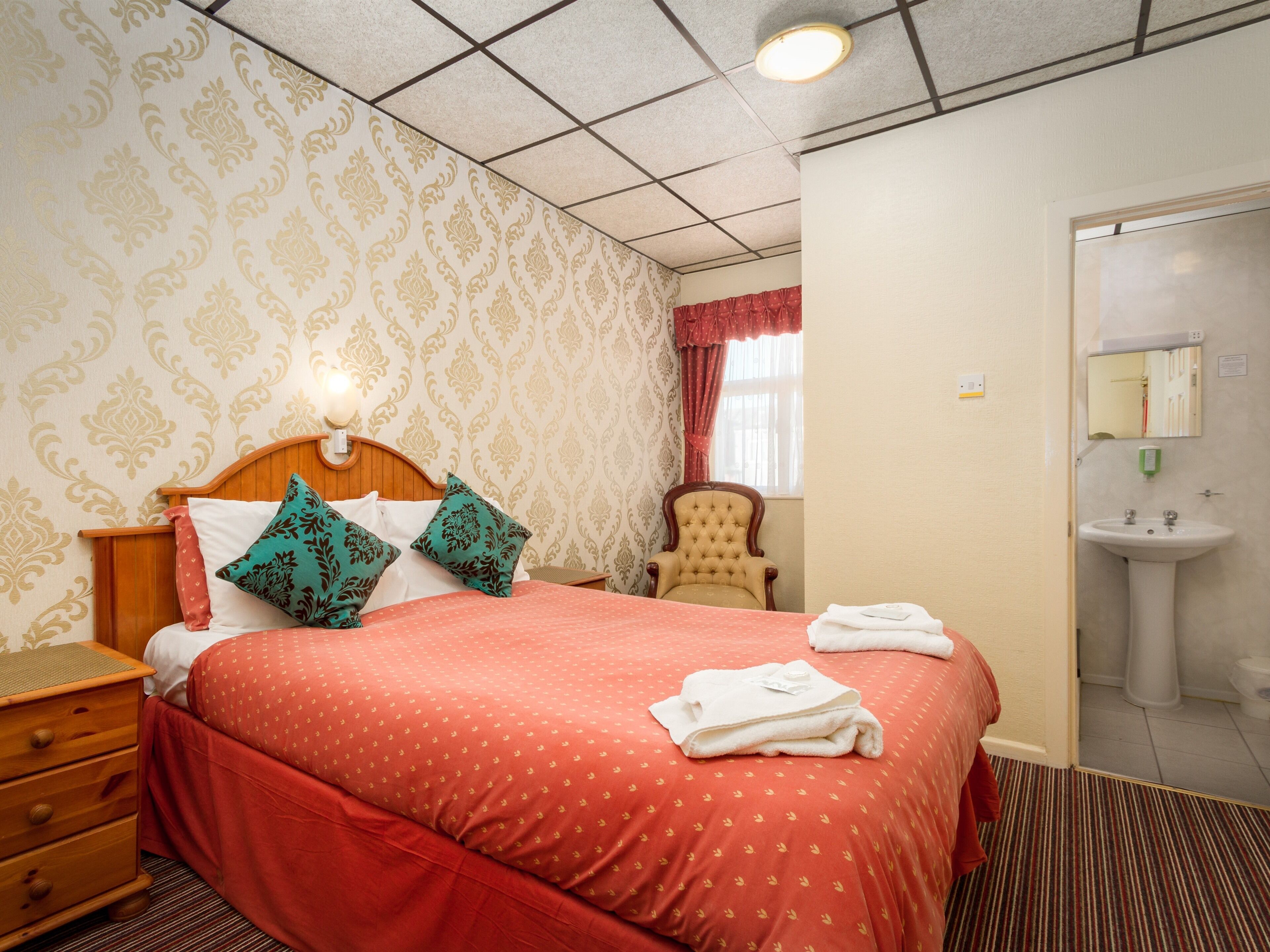 A hotel room with a large bed, a wooden nightstand, a chair, a bathroom with a sink and mirror, and patterned wallpaper.
