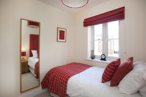 A bedroom with a bed, a window with a red valance, a bedside table with a lamp, a mirror, and a framed picture on the wall.