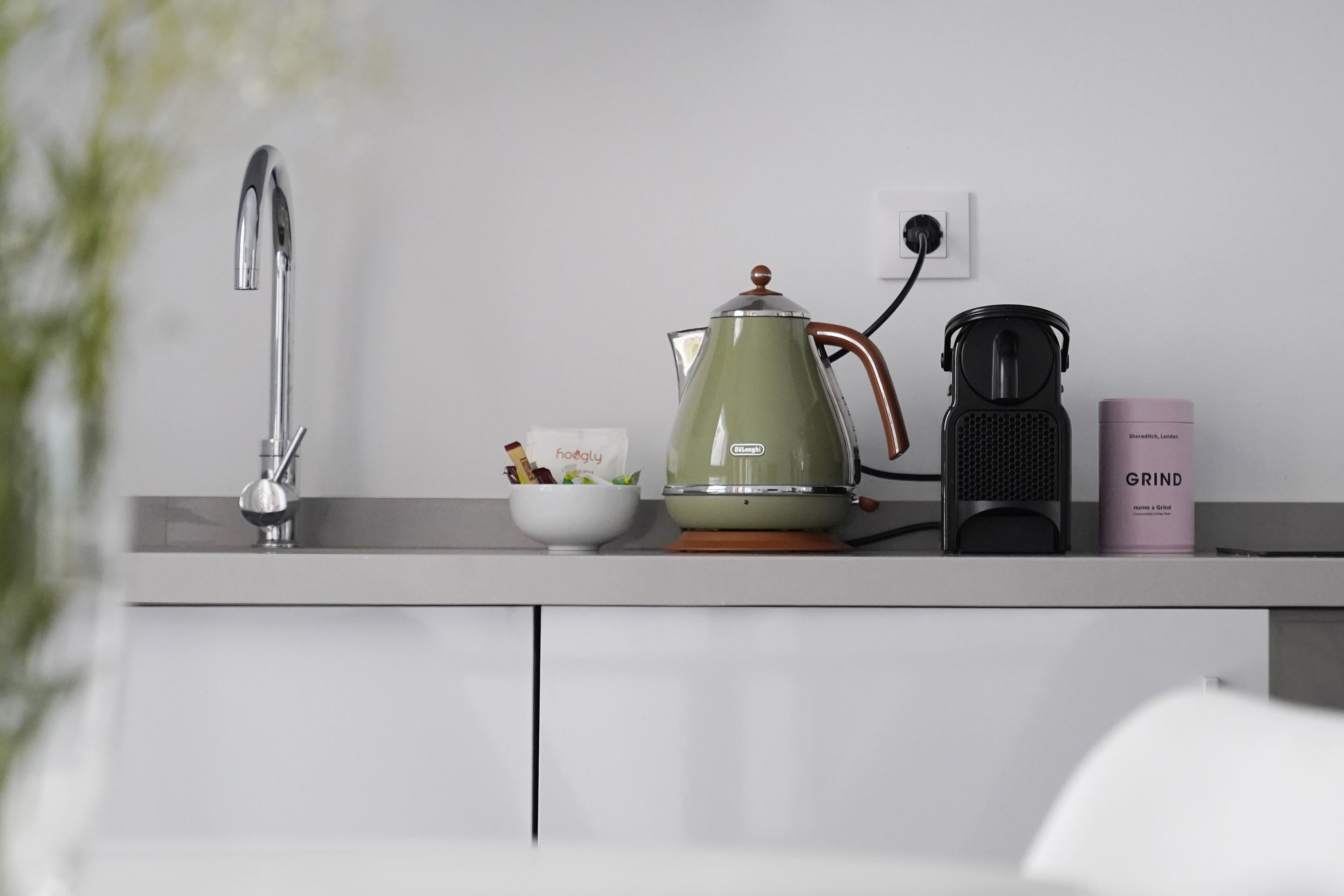 A kitchen counter with a kettle, a coffee maker, a bowl with food, and a pink container.