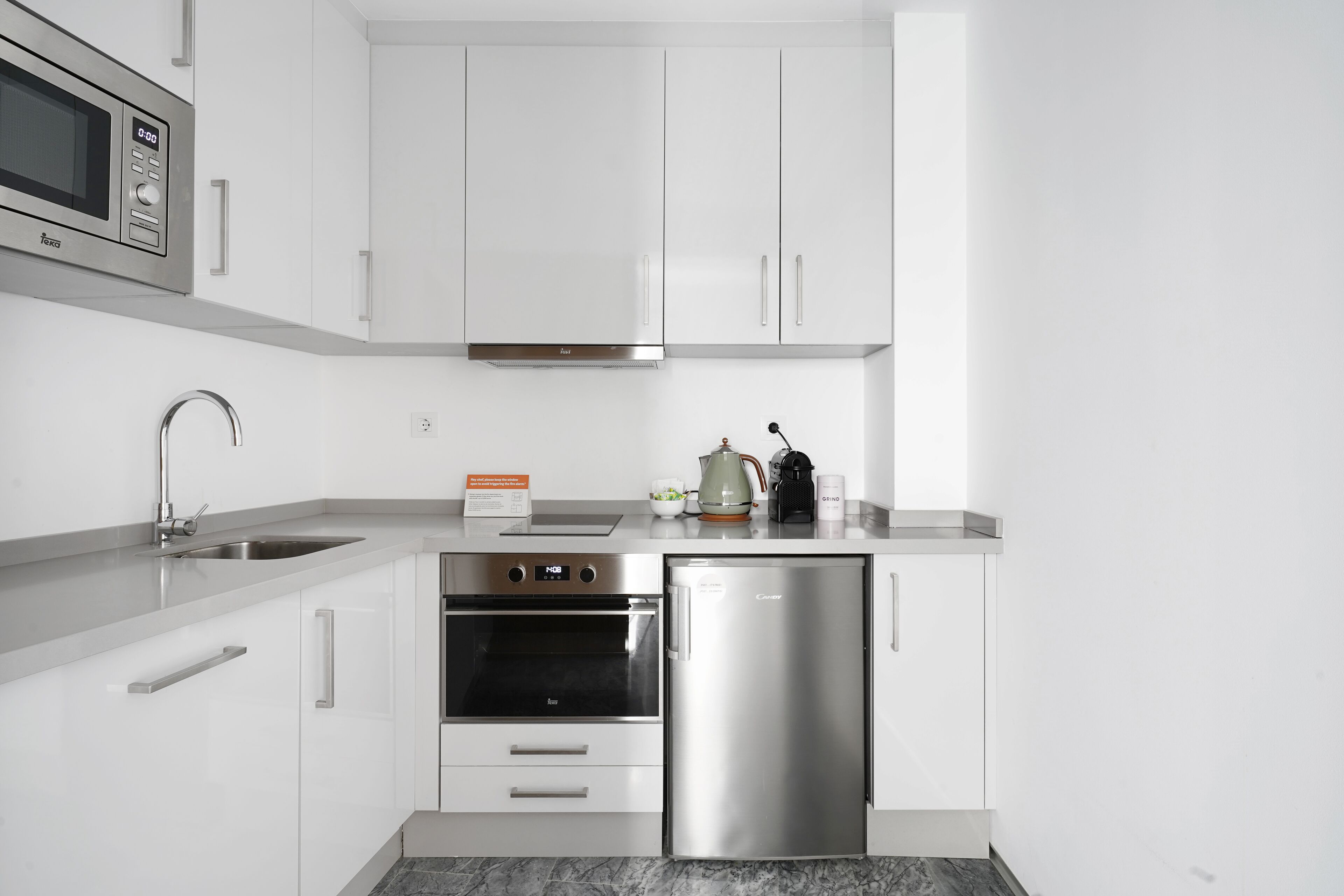 Een moderne keuken met witte kastjes, roestvrijstalen apparatuur, waaronder een magnetron, oven en koelkast, een spoelbak en een aanrecht met waterkoker en koffiezetapparaat.