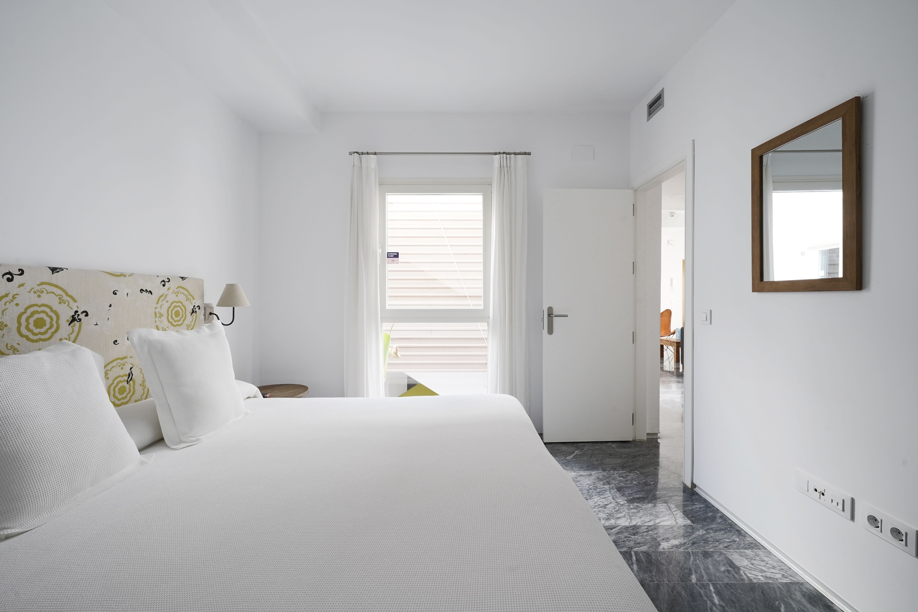 A hotel room with a bed, a nightstand, a mirror, and a door leading to another room.
