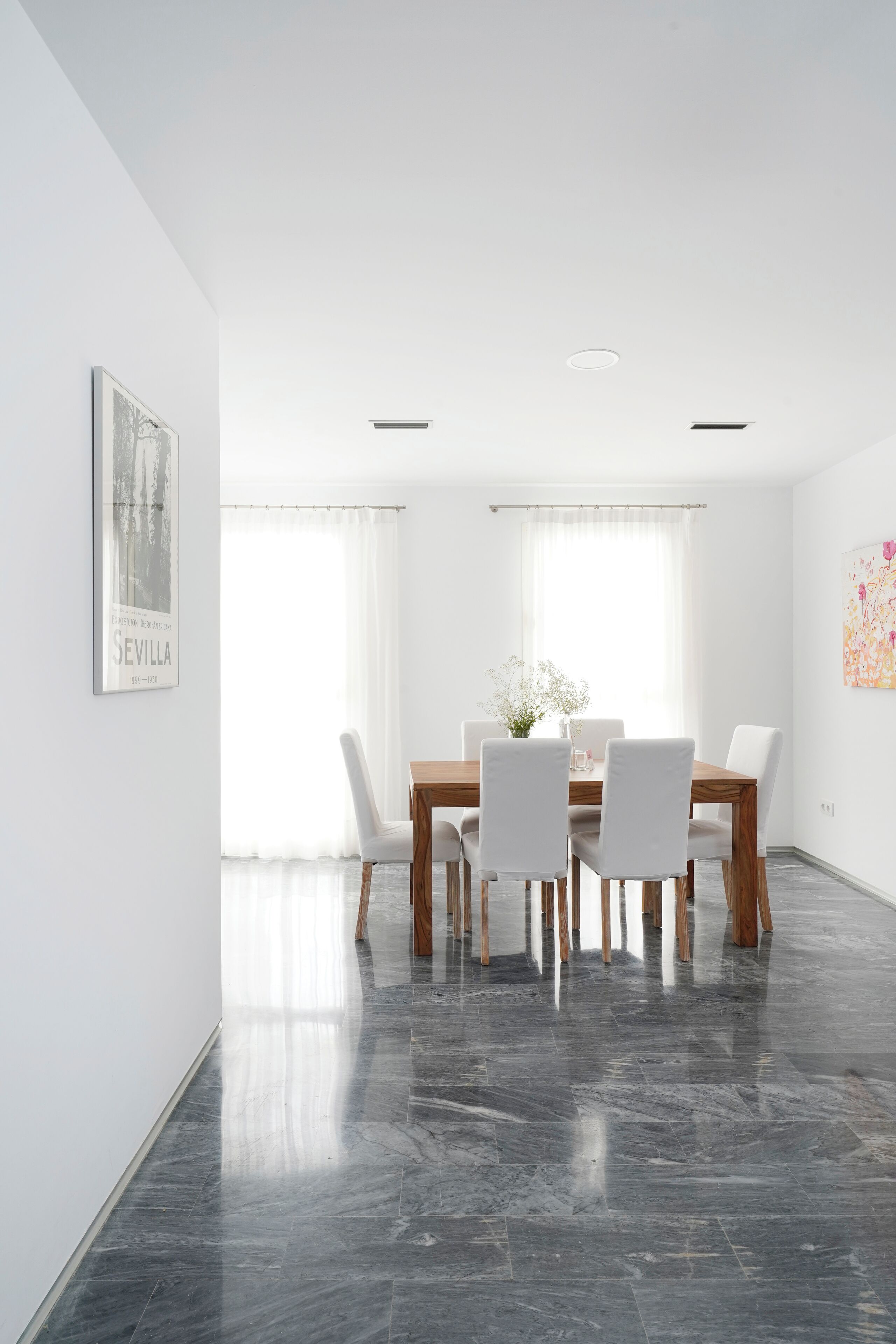 Un'area pranzo con tavolo in legno e sedie bianche, un quadro appeso alla parete e una finestra con tende leggere.