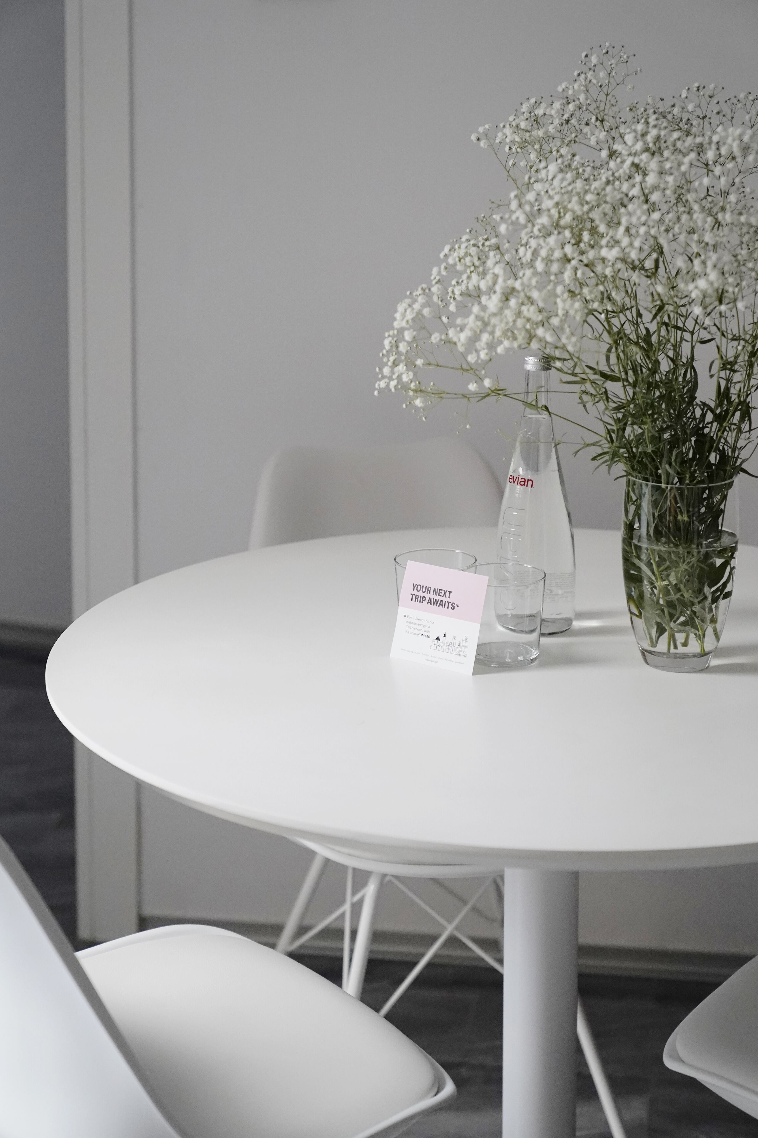 A round white table with a clear glass bottle, a glass of water, and a vase with white flowers. There is a sign on the table.