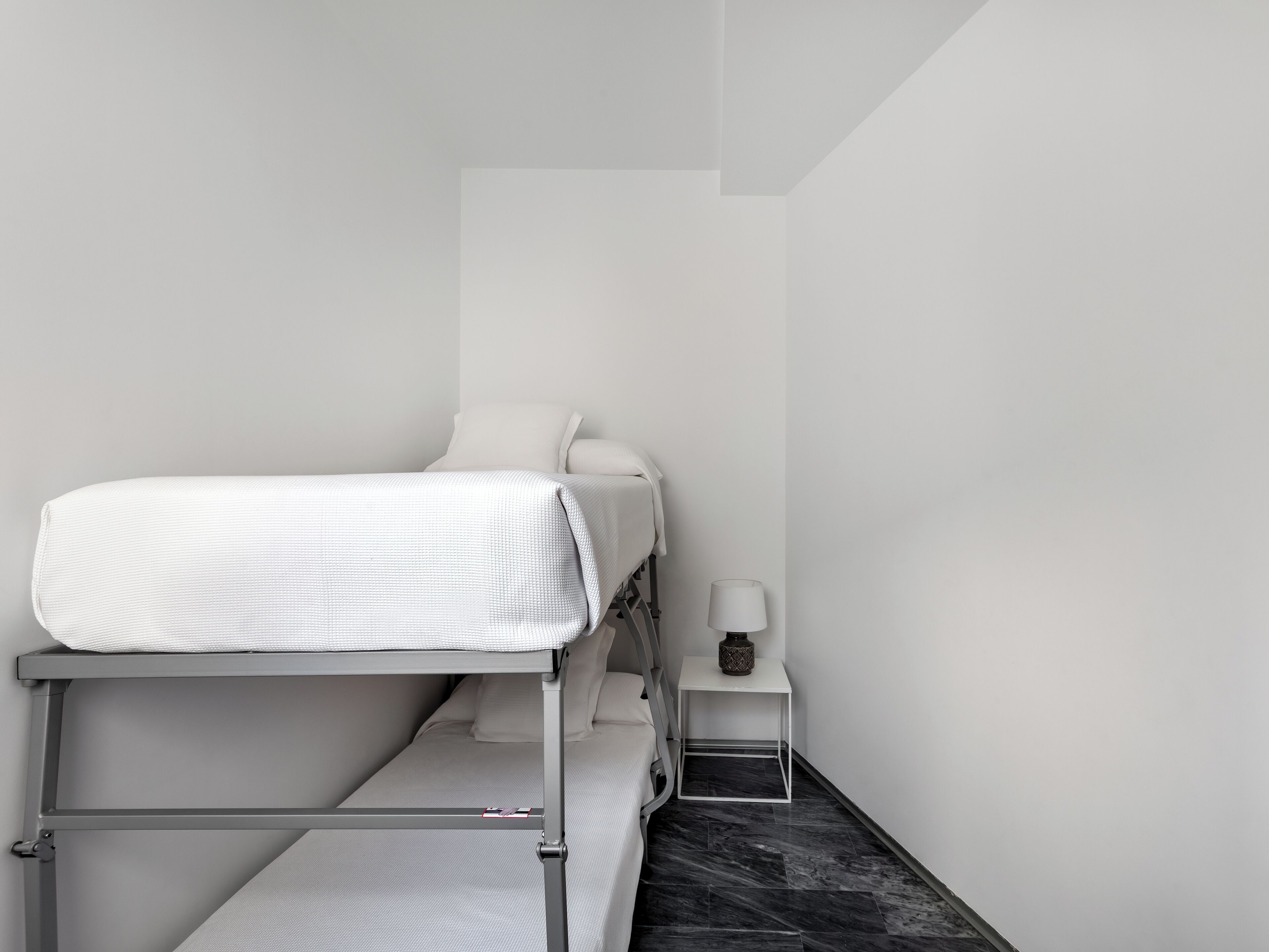 Habitación minimalista con literas, una mesita de noche pequeña con lámpara y suelo de mármol.