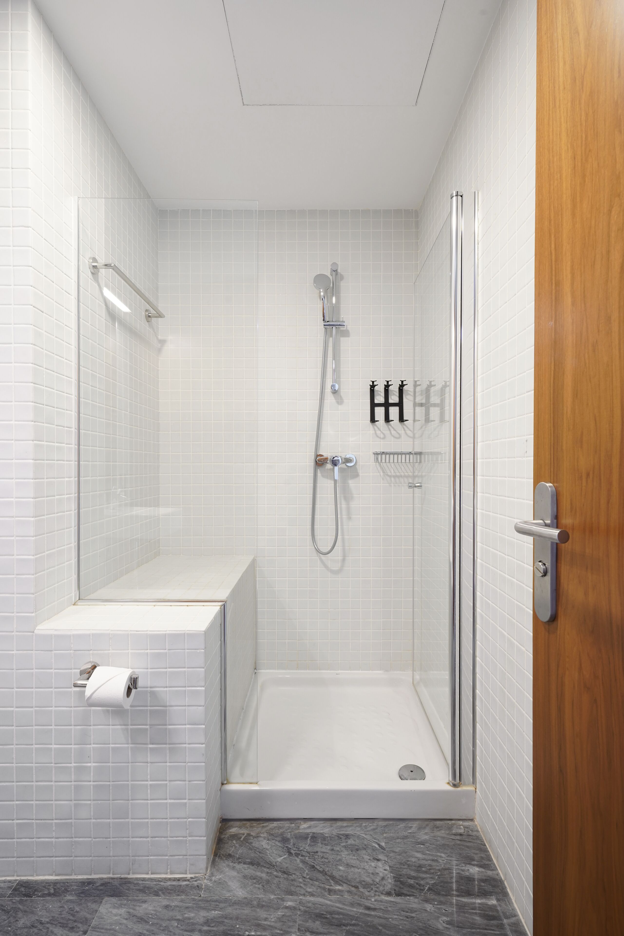 A modern shower room with a glass door, white tiled walls, a marble floor, and a wooden door.