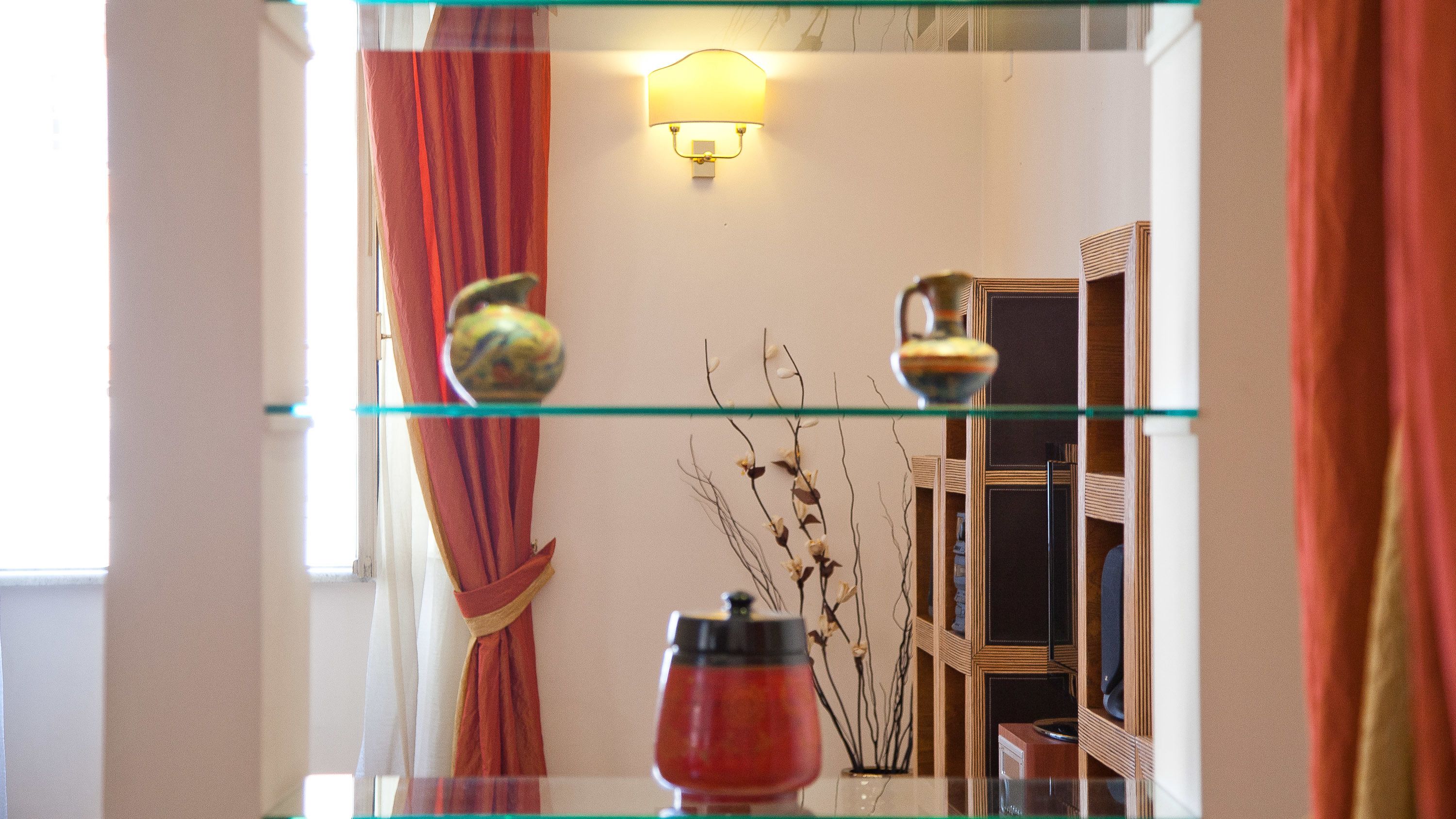 A room with a glass shelf displaying decorative items, a wall-mounted light fixture, and a mirror reflecting a window with curtains.