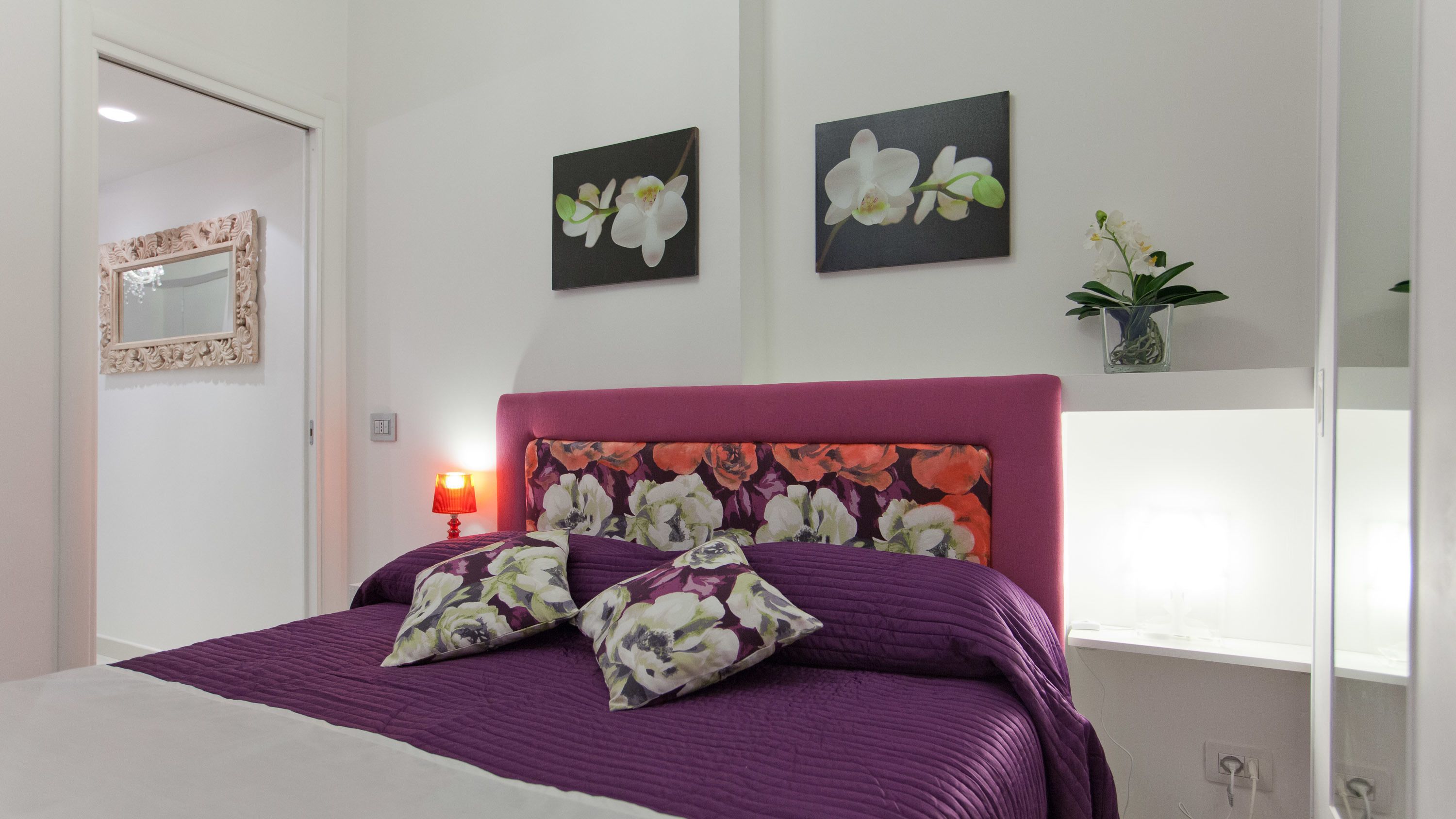 Un letto con una coperta viola e cuscini floreali, una testiera rossa e due dipinti di orchidee al muro.