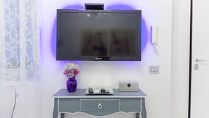 Televisão de tela plana fixada na parede, acima de uma mesa pequena com um vaso de flores, um controle remoto e uma caixa.