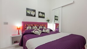 Um quarto com colcha roxa, almofadas a combinar, abajur vermelho e quadros com motivos florais nas paredes.