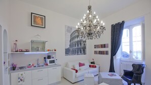 A modern living room with a white sofa, a dining table, a chandelier, and a large window with curtains.