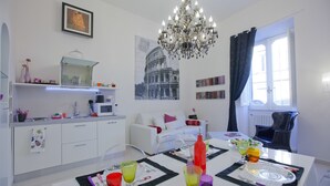 A modern living room with a dining area, a chandelier, a kitchen with a microwave, and a large window with curtains.