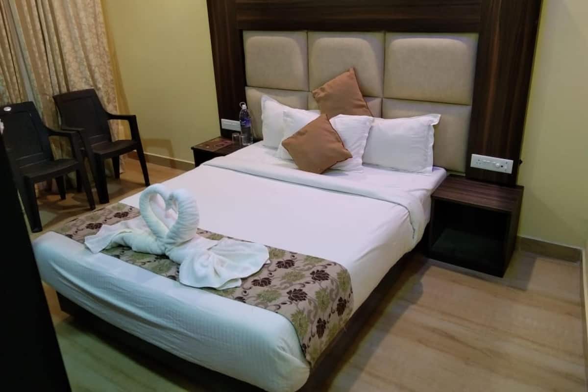 A neatly made bed with a floral bedspread, a wooden headboard, and a set of pillows. There is a small table with a water bottle and a remote control. A chair is visible in the corner.