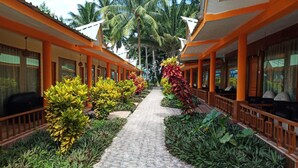 Un pasillo con piso de baldosas, bordeado por hileras de habitaciones con paredes anaranjadas y blancas, y un camino central bordeado de plantas tropicales.