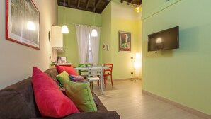 A cozy living room with a brown sofa, colorful cushions, a dining area with a table and chairs, a flat-screen TV mounted on the wall, and a kitchen area with a stove and sink. - Rental in Rome Crociferi 2 (Rome)