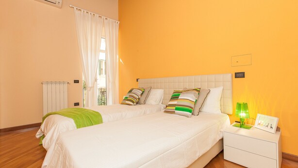 A bedroom with a large bed, white bedding, a green throw blanket, and striped pillows. There is a nightstand with a lamp and a framed picture. - Rental in Rome Crociferi 2 (Rome)