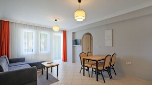 A modern living room with a sofa, a dining table with chairs, a TV mounted on the wall, and a large window with curtains. - Gold Lion Apart Hotel (Fethiye)
