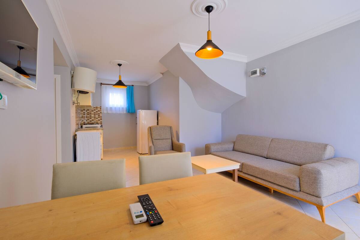 A modern living room with a sofa, a dining table with a remote control, and a kitchen in the background.