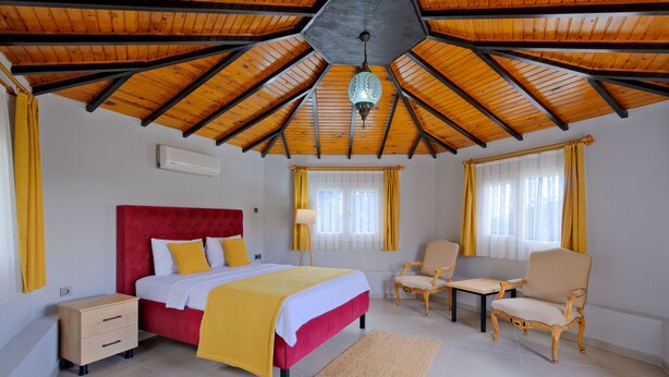 A bedroom with a wooden ceiling, a bed with a red headboard, a bedside table, a chair, and a small table. - Gold Lion Apart Hotel (Fethiye)