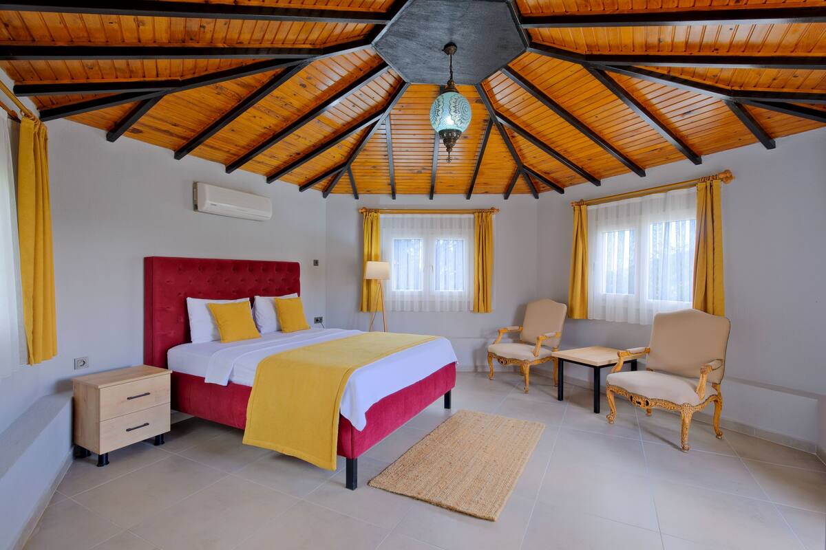A bedroom with a wooden ceiling, a bed with a red headboard, a bedside table, a chair, and a small table.