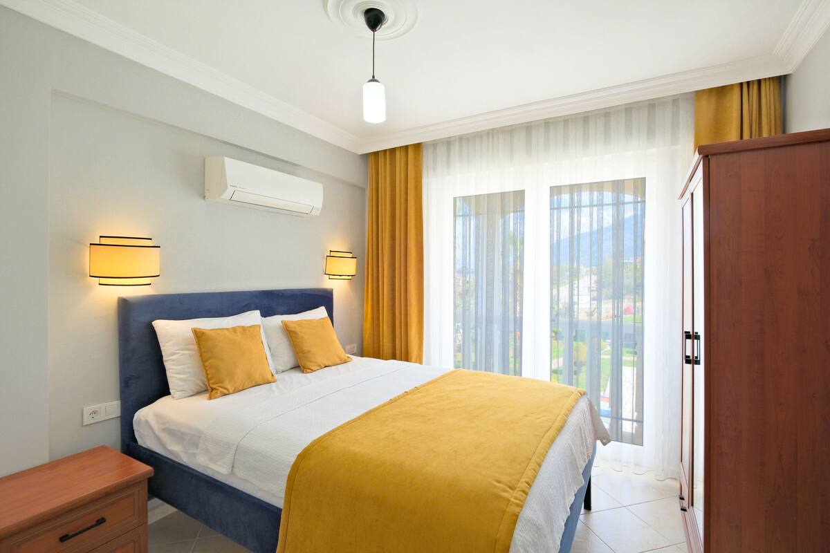 A hotel room with a bed, bedside lamps, a wardrobe, and a view of the outdoors through a large window.