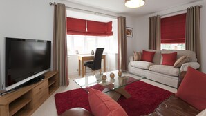 A modern living room with a flat-screen TV, a glass coffee table, a sofa, and a dining area with a chair and table. - The Mews (Derby)