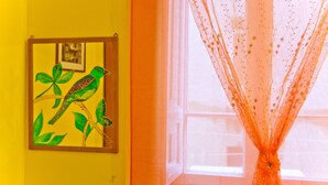 A room with a yellow wall, a framed artwork of a bird on a branch, and a window with sheer curtains.