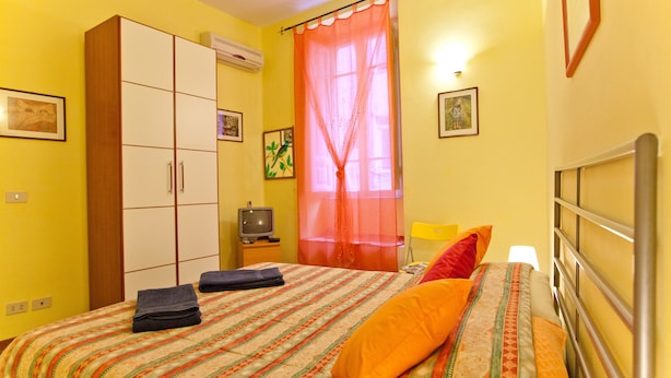 A bedroom with a bed, a wardrobe, a television, and a window with a sheer curtain.